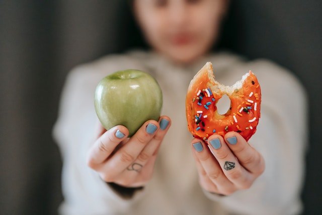 A girl holding an apple and a doughnut portraying one of the reasons why you're not seeing results from working out.
